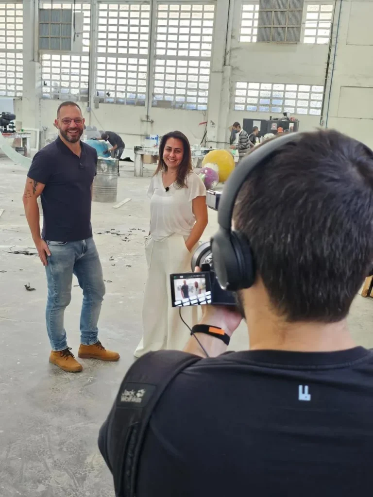 Filmagem em fábrica com dois apresentadores e câmera.